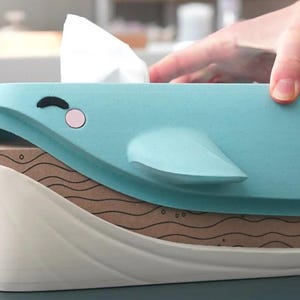 May include: A light blue whale-shaped tissue box cover with a white base. The whale has a black eye and a pink cheek. The tissue box is sitting on top of a brown box with a wavy pattern.