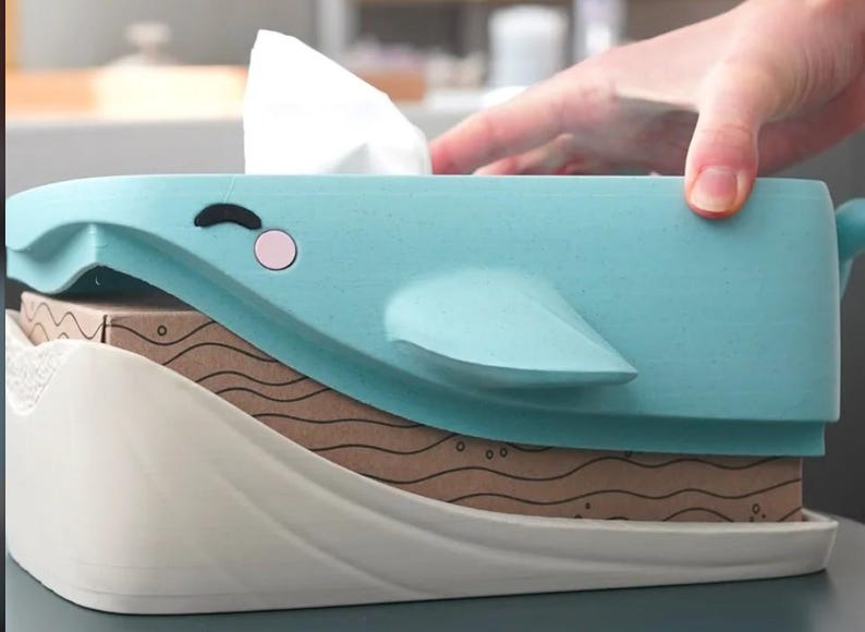May include: A light blue whale-shaped tissue box cover with a white base. The whale has a black eye and a pink cheek. The tissue box is sitting on top of a brown box with a wavy pattern.