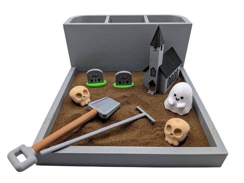 Pu&ograve; includere: Un organizzatore da scrivania in legno grigio a tema Halloween. Presenta una piccola chiesa, un fantasma, due lapidi, due teschi, una pala e un rastrello. L'organizzatore &egrave; riempito di sabbia marrone.