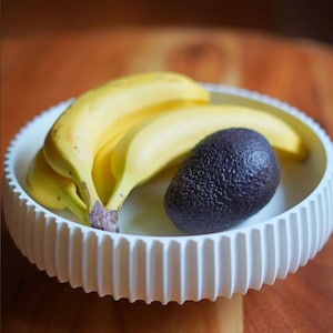 Puede incluir: Un cuenco de cer&aacute;mica blanco y acanalado contiene un racimo de pl&aacute;tanos amarillos maduros y un aguacate morado oscuro. El cuenco est&aacute; sobre una superficie de madera, una naturaleza muerta sencilla.
