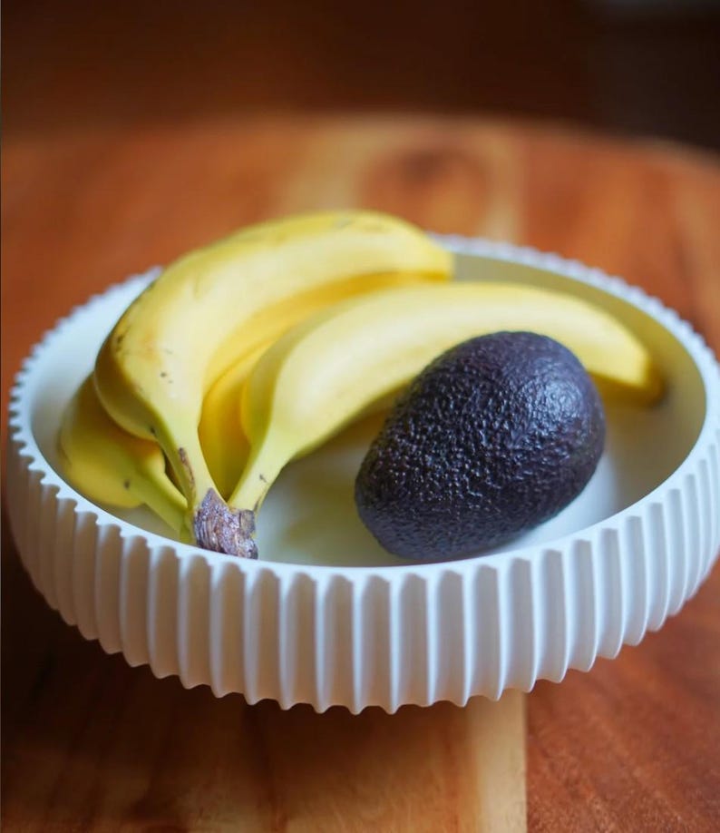 Puede incluir: Un cuenco de cer&aacute;mica blanco y acanalado contiene un racimo de pl&aacute;tanos amarillos maduros y un aguacate morado oscuro. El cuenco est&aacute; sobre una superficie de madera, una naturaleza muerta sencilla.