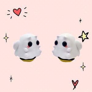 May include: Two white ghost-shaped figurines with black eyes and pink cheeks, each with a yellow and black bee-shaped base. The figurines are facing each other on a pink background.