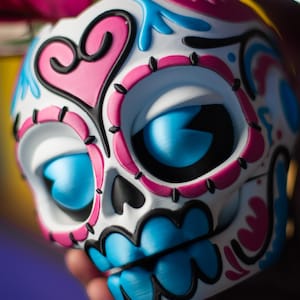 May include: A decorative Day of the Dead skull with a white base, adorned with pink, blue, and black accents. The skull features large blue eyes, a pink heart, and intricate floral designs, held in a hand.