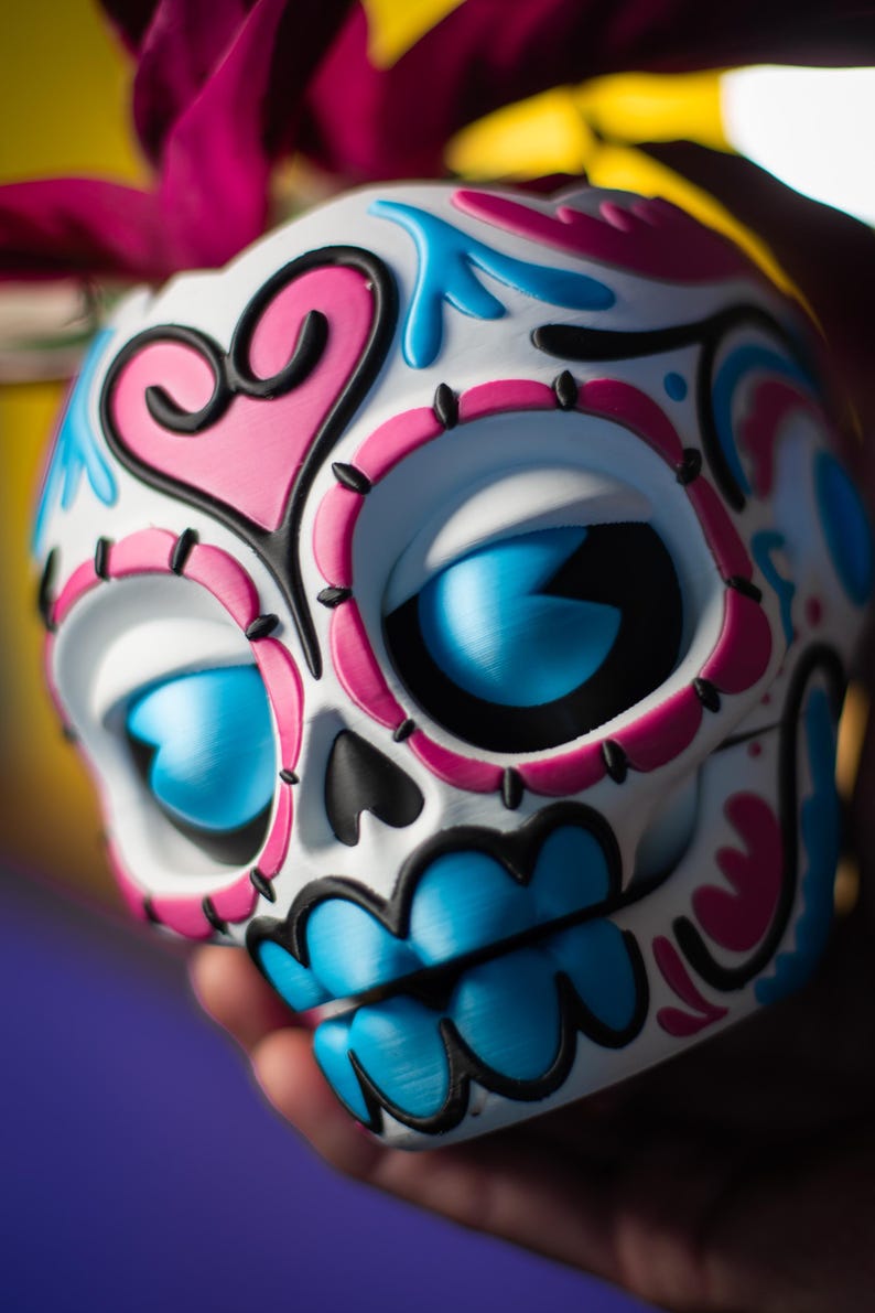 May include: A decorative Day of the Dead skull with a white base, adorned with pink, blue, and black accents. The skull features large blue eyes, a pink heart, and intricate floral designs, held in a hand.