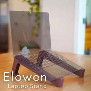 May include: A purple laptop stand supporting a laptop. The stand is angled, with the text "Elowen Laptop Stand" in white. The laptop is open, showing the keyboard and screen.