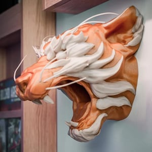 May include: A 3D-printed tiger head sculpture in shades of orange and white, mounted on a wall. The tiger's mouth is open, revealing sharp teeth. The sculpture has detailed fur and whisker elements, creating a realistic appearance.