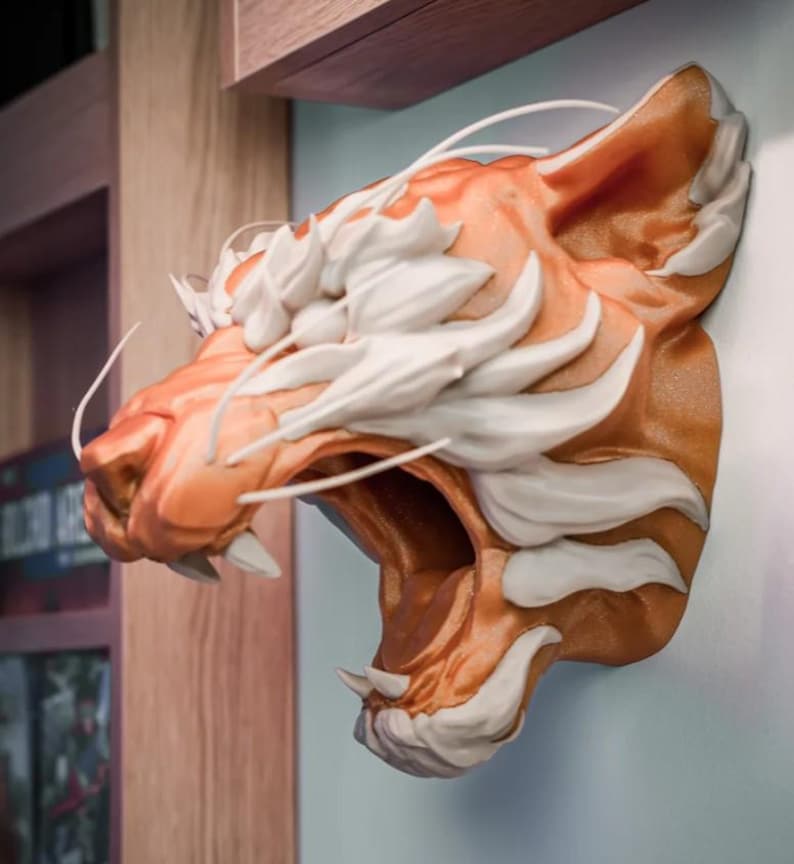 May include: A 3D-printed tiger head sculpture in shades of orange and white, mounted on a wall. The tiger's mouth is open, revealing sharp teeth. The sculpture has detailed fur and whisker elements, creating a realistic appearance.