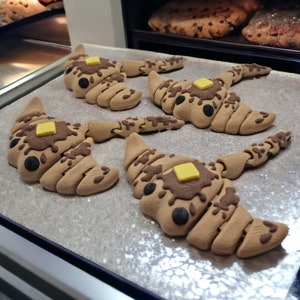 Könnte beinhalten: Vier braune, croissantförmige Backwaren mit Schokoladenguss und einem Quadrat aus gelber Butter obenauf. Die Backwaren sind auf einem Backpapier in einem Backgerät angeordnet. Weitere Backwaren sind im Hintergrund zu sehen.