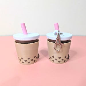May include: Two miniature boba tea cup keychains. The cups are tan with brown boba pearls and a white lid. Each has a pink straw and one has a rose gold clasp. The background is a solid pink.