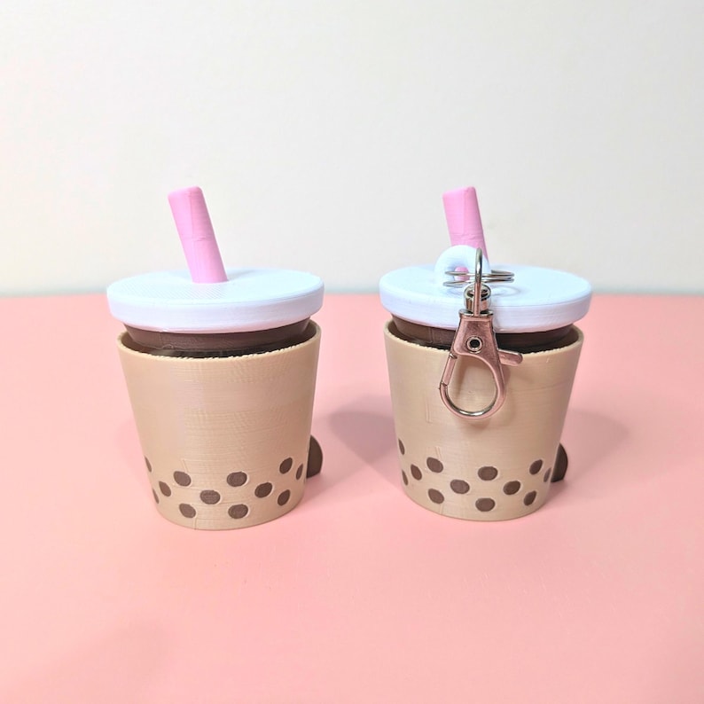 May include: Two miniature boba tea cup keychains. The cups are tan with brown boba pearls and a white lid. Each has a pink straw and one has a rose gold clasp. The background is a solid pink.