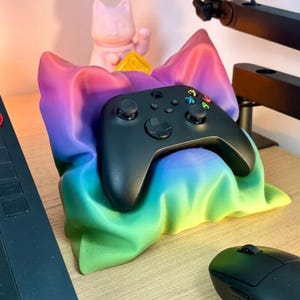 May include: A black Xbox controller resting in a rainbow-colored pillow. The pillow is made of a soft, plush fabric and has a gradient of colors from purple to green. The controller is sitting on a wooden surface.
