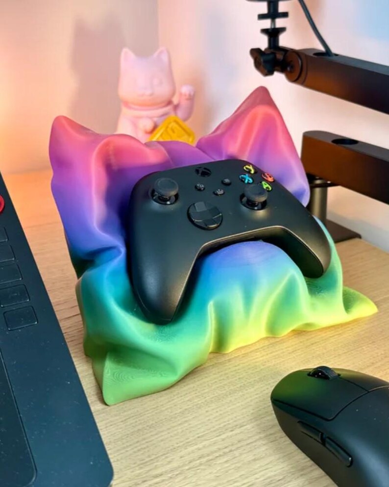 May include: A black Xbox controller resting in a rainbow-colored pillow. The pillow is made of a soft, plush fabric and has a gradient of colors from purple to green. The controller is sitting on a wooden surface.