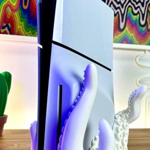 May include: A white 3D printed octopus stand for a video game console. The stand has two tentacles on the left and one on the right. The console is white and black and is held in place by the tentacles.