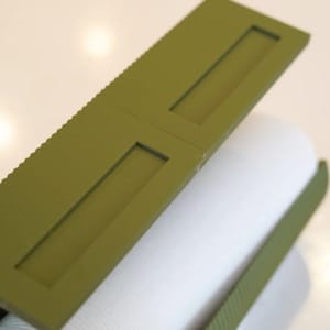 May include: Olive green rectangular object with two rectangular cutouts, resting on a white cylindrical object. The top edge of the green object has a ridged texture. The objects are on a white surface.
