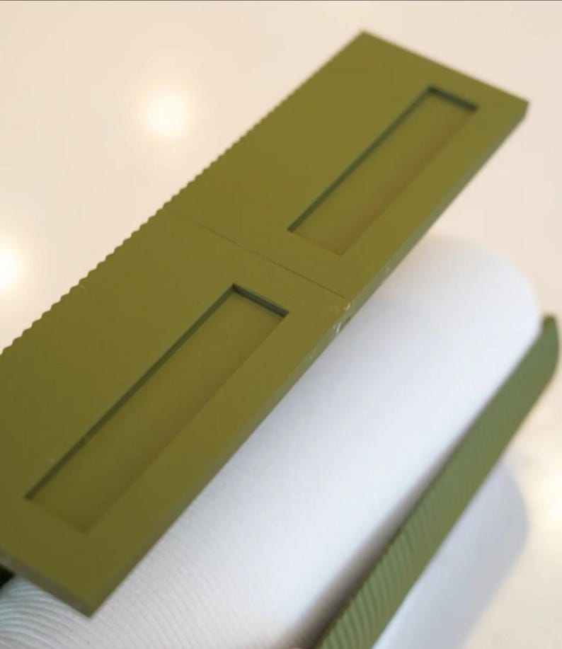 May include: Olive green rectangular object with two rectangular cutouts, resting on a white cylindrical object. The top edge of the green object has a ridged texture. The objects are on a white surface.