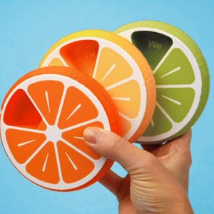 Puede incluir: Tres accesorios de cocina con forma de fruta se sostienen en una mano sobre un fondo azul. Los accesorios se asemejan a rodajas de naranja, limón y lima, con detalles blancos. La rodaja de lima tiene la palabra "We".
