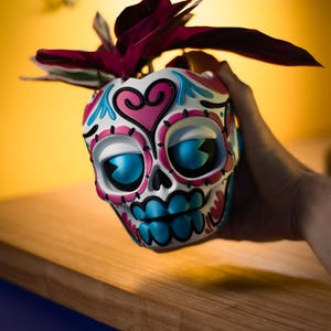 May include: A decorative skull-shaped planter with a white base, adorned with pink, blue, and black details. The planter features large blue eyes and a heart design. It holds a plant with dark red leaves, creating a unique decorative piece.