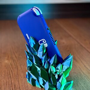May include: A blue Nintendo Switch Lite console is standing upright in a green and blue crystal-shaped stand. The stand is made of 3D printed plastic and has a textured, crystalline appearance.