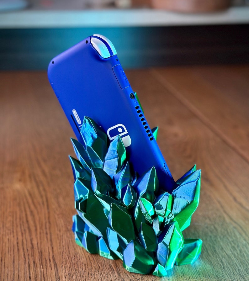 May include: A blue Nintendo Switch Lite console is standing upright in a green and blue crystal-shaped stand. The stand is made of 3D printed plastic and has a textured, crystalline appearance.