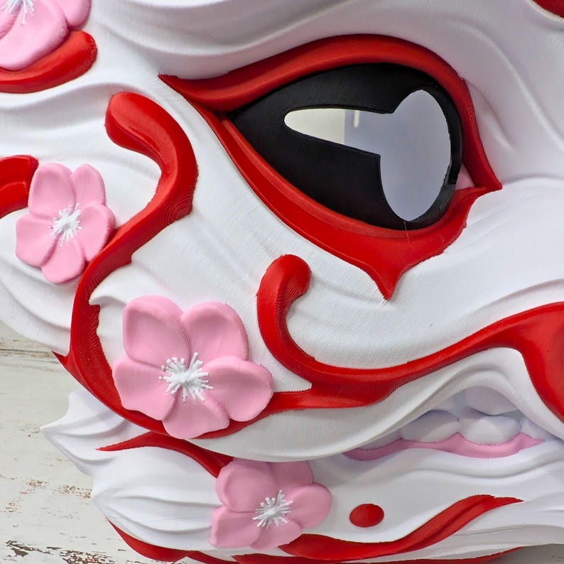 May include: Close-up of a white and red decorative mask with a black eye and pink flower accents. The mask has a detailed design with curved lines and a textured surface, resembling a mythical creature.