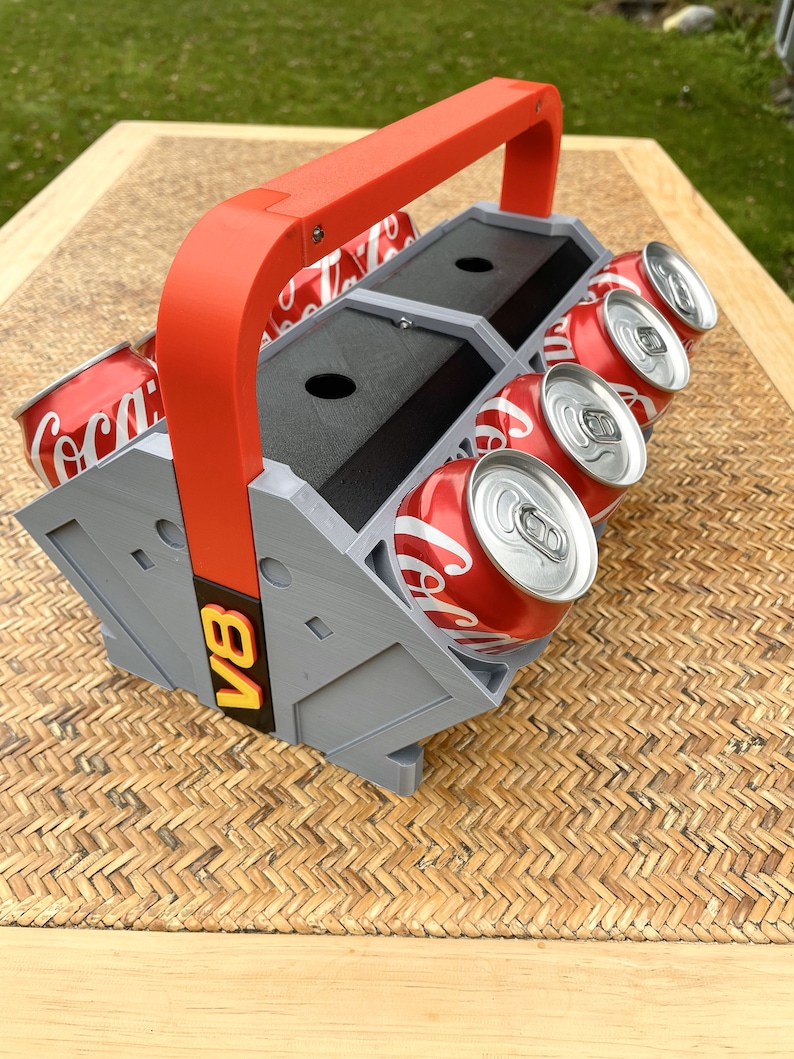 May include: A red and gray 3D printed can holder with a handle. The holder has the text "V8" on the side and can hold six cans. The cans are red and white and have the Coca-Cola logo.