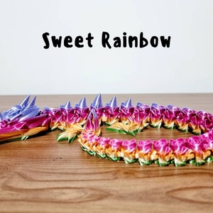Peut inclure: Un dragon color&eacute;, aux couleurs de l'arc-en-ciel, fait de papier origami. Le dragon est enroul&eacute; sur une surface en bois. Le texte "Sweet Rainbow" est &eacute;crit au-dessus du dragon.