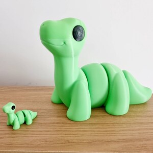 Puede incluir: Dos juguetes de dinosaurio impresos en 3D de color verde. El dinosaurio m&aacute;s grande est&aacute; de pie sobre sus patas traseras con la cola enroscada alrededor de su cuerpo. El dinosaurio m&aacute;s peque&ntilde;o est&aacute; acostado sobre su vientre con la cabeza levantada. Ambos dinosaurios tienen ojos negros.