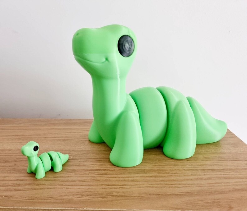 Puede incluir: Dos juguetes de dinosaurio impresos en 3D de color verde. El dinosaurio m&aacute;s grande est&aacute; de pie sobre sus patas traseras con la cola enroscada alrededor de su cuerpo. El dinosaurio m&aacute;s peque&ntilde;o est&aacute; acostado sobre su vientre con la cabeza levantada. Ambos dinosaurios tienen ojos negros.
