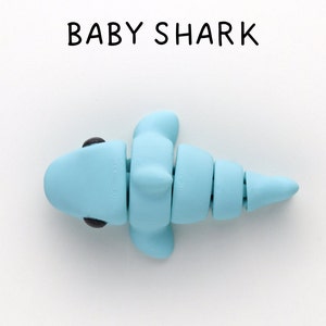 May include: A light blue baby shark made of clay with black eyes. The shark is facing left and has a segmented tail.
