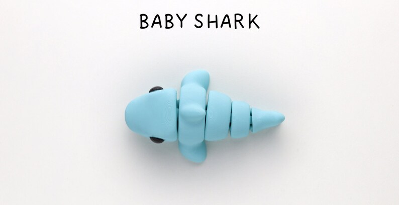 May include: A light blue baby shark made of clay with black eyes. The shark is facing left and has a segmented tail.