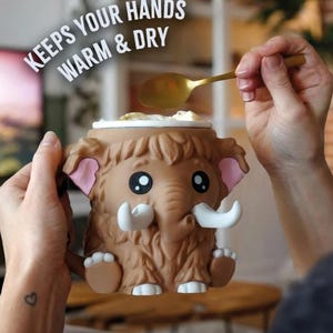 May include: A brown mammoth-shaped mug with large eyes, white tusks, and pink ears. The mug has the text "KEEPS YOUR HANDS WARM & DRY" in white. A gold spoon is scooping from the top of the mug.