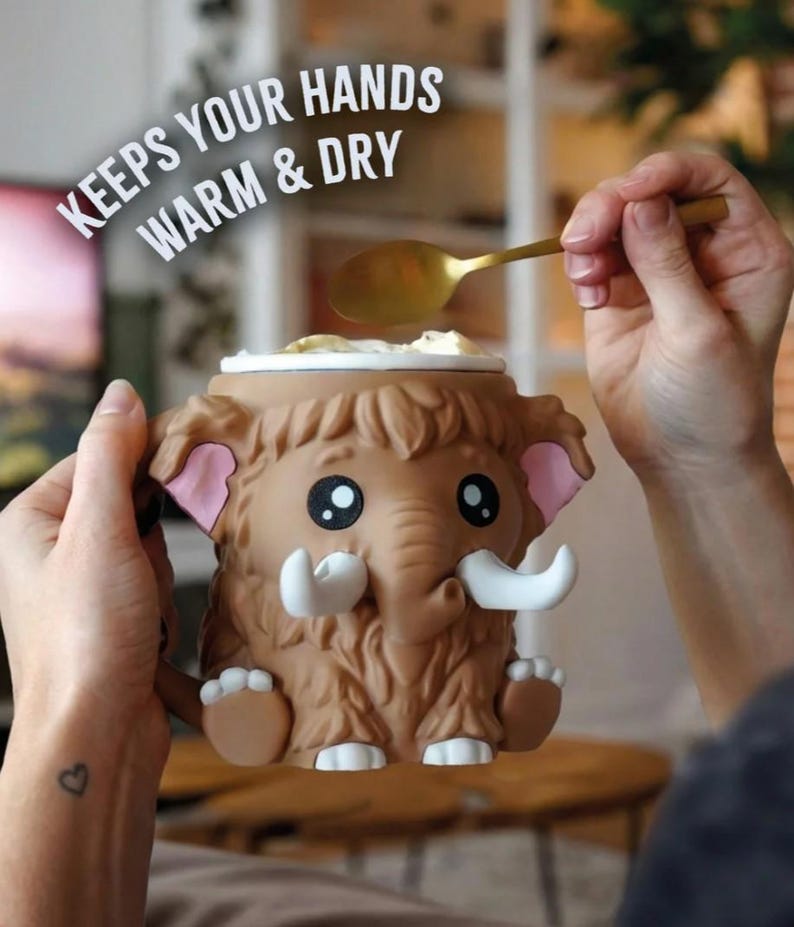 May include: A brown mammoth-shaped mug with large eyes, white tusks, and pink ears. The mug has the text "KEEPS YOUR HANDS WARM & DRY" in white. A gold spoon is scooping from the top of the mug.