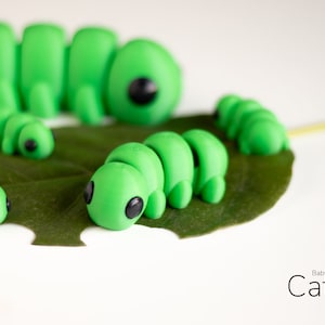 Puede incluir: Varias figuras de orugas de color verde brillante con ojos negros descansan sobre una hoja verde. Las orugas var&iacute;an en tama&ntilde;o, siendo la m&aacute;s grande la que est&aacute; en el fondo. El texto "Baby Caterpillar" est&aacute; en la esquina inferior derecha.