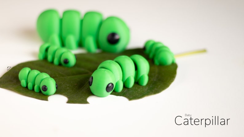 Puede incluir: Varias figuras de orugas de color verde brillante con ojos negros descansan sobre una hoja verde. Las orugas var&iacute;an en tama&ntilde;o, siendo la m&aacute;s grande la que est&aacute; en el fondo. El texto "Baby Caterpillar" est&aacute; en la esquina inferior derecha.