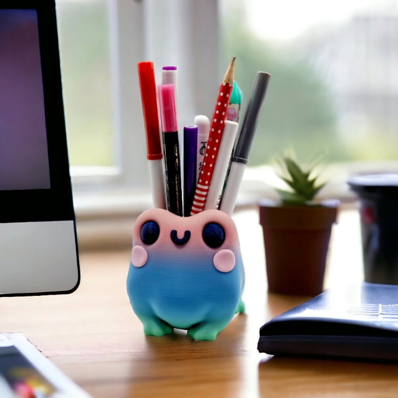 Pu&ograve; includere: Un portapenne stravagante a forma di rana, stampato in 3D con una sfumatura di rosa, blu e verde. La rana ha grandi occhi blu e una bocca sorridente. Contiene penne e matite, aggiungendo un tocco giocoso a una scrivania o spazio di lavoro.
