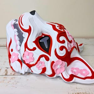 May include: A white, red, and pink decorative mask in the shape of a wolf's head. The mask features red swirling patterns, a black eye, and pink flower accents. The wolf's mouth is open, revealing pink and gray details.