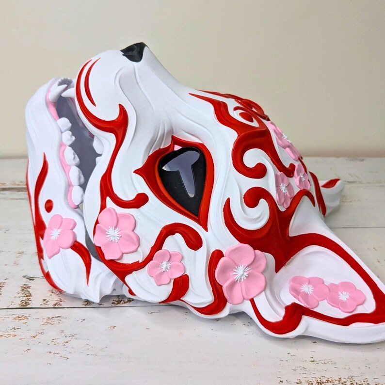 May include: A white, red, and pink decorative mask in the shape of a wolf's head. The mask features red swirling patterns, a black eye, and pink flower accents. The wolf's mouth is open, revealing pink and gray details.