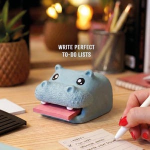 May include: A light blue hippo-shaped sticky note holder with a pink pad of sticky notes. The text "WRITE PERFECT TO-DO LISTS" is visible. A person is writing on a to-do list.