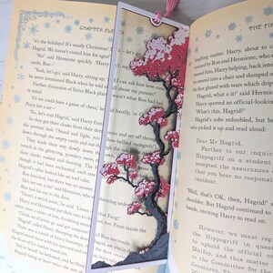 May include: A handmade bookmark featuring a detailed tree design with red and white blossoms. The bookmark is set against the pages of a book, with a pink tassel attached at the top.