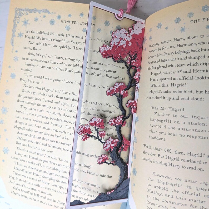 May include: A handmade bookmark featuring a detailed tree design with red and white blossoms. The bookmark is set against the pages of a book, with a pink tassel attached at the top.