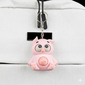 May include: A pink pig-shaped keychain with large, expressive eyes and a round belly button design. The keychain is attached to a silver keyring and black strap, hanging against a white textured background.