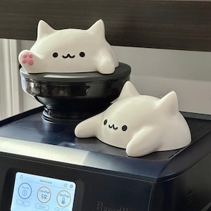May include: Two white cat-shaped figurines with black eyes and a smiling mouth. One cat has a pink paw. The figurines are on top of a black coffee machine and a black grinder. The coffee machine has a digital display.