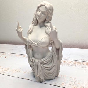 May include: A matte white sculpture of a figure with long flowing hair, wearing a draped top and skirt, depicted from the waist up. The figure has both arms raised with the middle fingers extended in a gesture, showcasing detailed fabric folds.