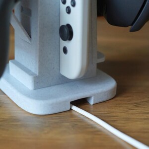 May include: A light gray gaming console stand with a white controller and a black headset. The stand has a slot for a charging cable. The stand is made of a speckled material and sits on a wooden surface.
