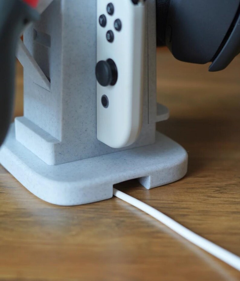 May include: A light gray gaming console stand with a white controller and a black headset. The stand has a slot for a charging cable. The stand is made of a speckled material and sits on a wooden surface.