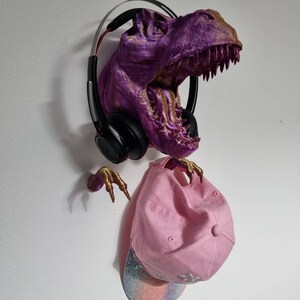 May include: A purple and gold dinosaur head wall mount with a pink baseball cap and black headphones hanging from it.