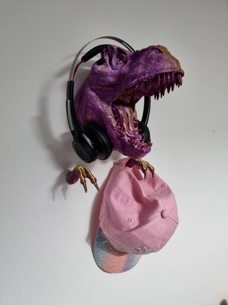 May include: A purple and gold dinosaur head wall mount with a pink baseball cap and black headphones hanging from it.