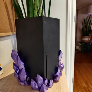 Puede incluir: Una consola de juegos negra con un soporte en forma de cristal morado. La consola est&aacute; sentada sobre una superficie de madera.