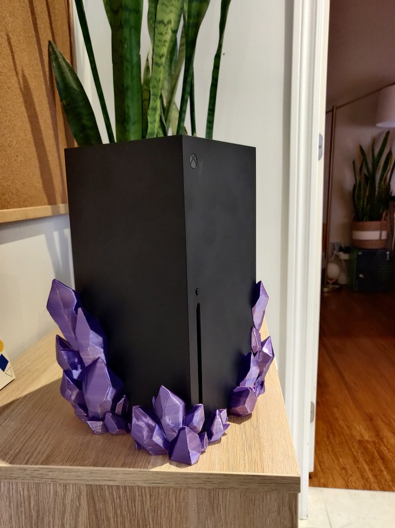 Puede incluir: Una consola de juegos negra con un soporte en forma de cristal morado. La consola est&aacute; sentada sobre una superficie de madera.