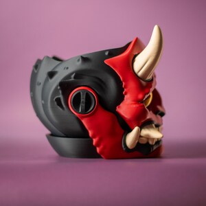 May include: A black and red decorative planter shaped like an Oni mask. The planter features a detailed design with horns, teeth, and a menacing expression. The planter is made of a durable material and is suitable for small plants.